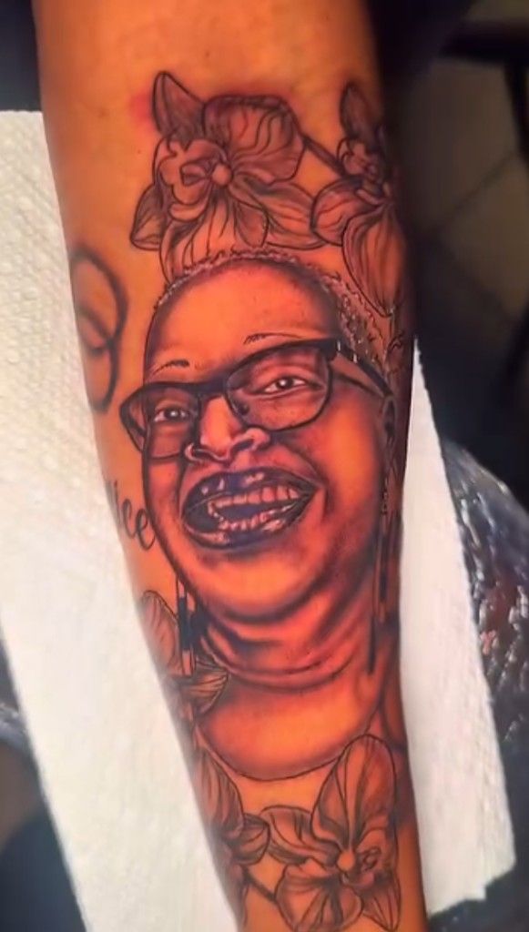 Black and grey portrait tattoo of a smiling person wearing glasses, framed by flowers.