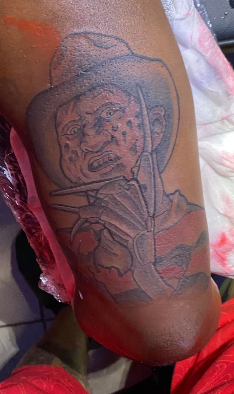 Portrait tattoo of a horror movie villain wearing a fedora and striped sweater on an upper arm.