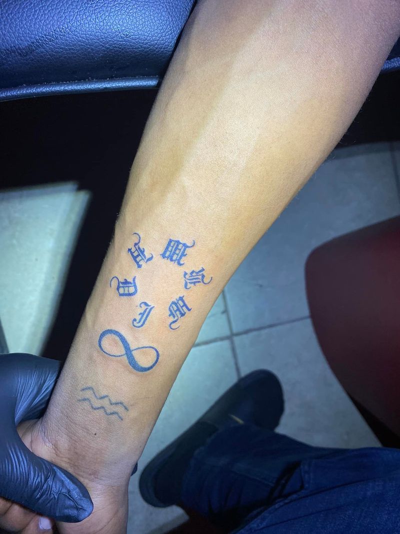 Small tattoos of old English letters, an infinity symbol and a wave symbol on a forearm.