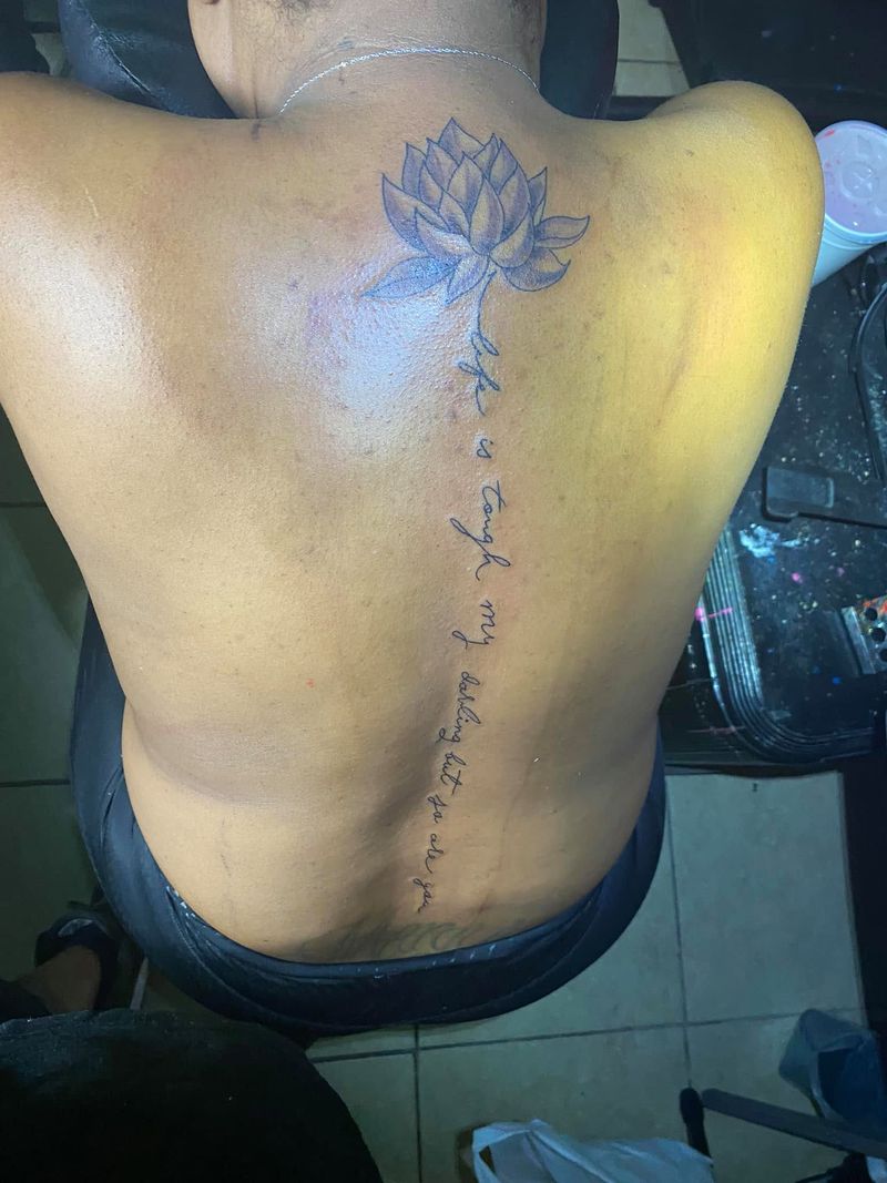 Tattoo of a lotus flower at the top of the spine with a line of script running down the back.