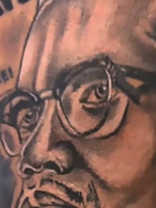 Close-up of a black and grey portrait tattoo of a man wearing round glasses.