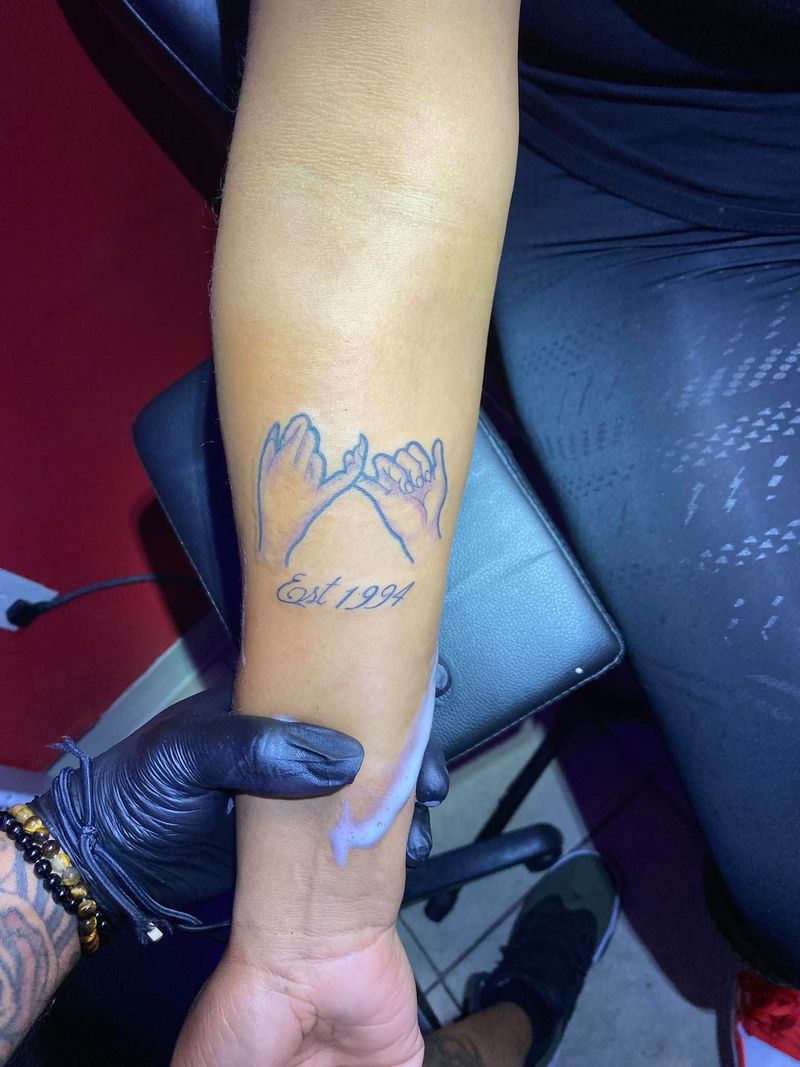 Fine line tattoo of two hands linking pinky fingers above small script on a forearm.