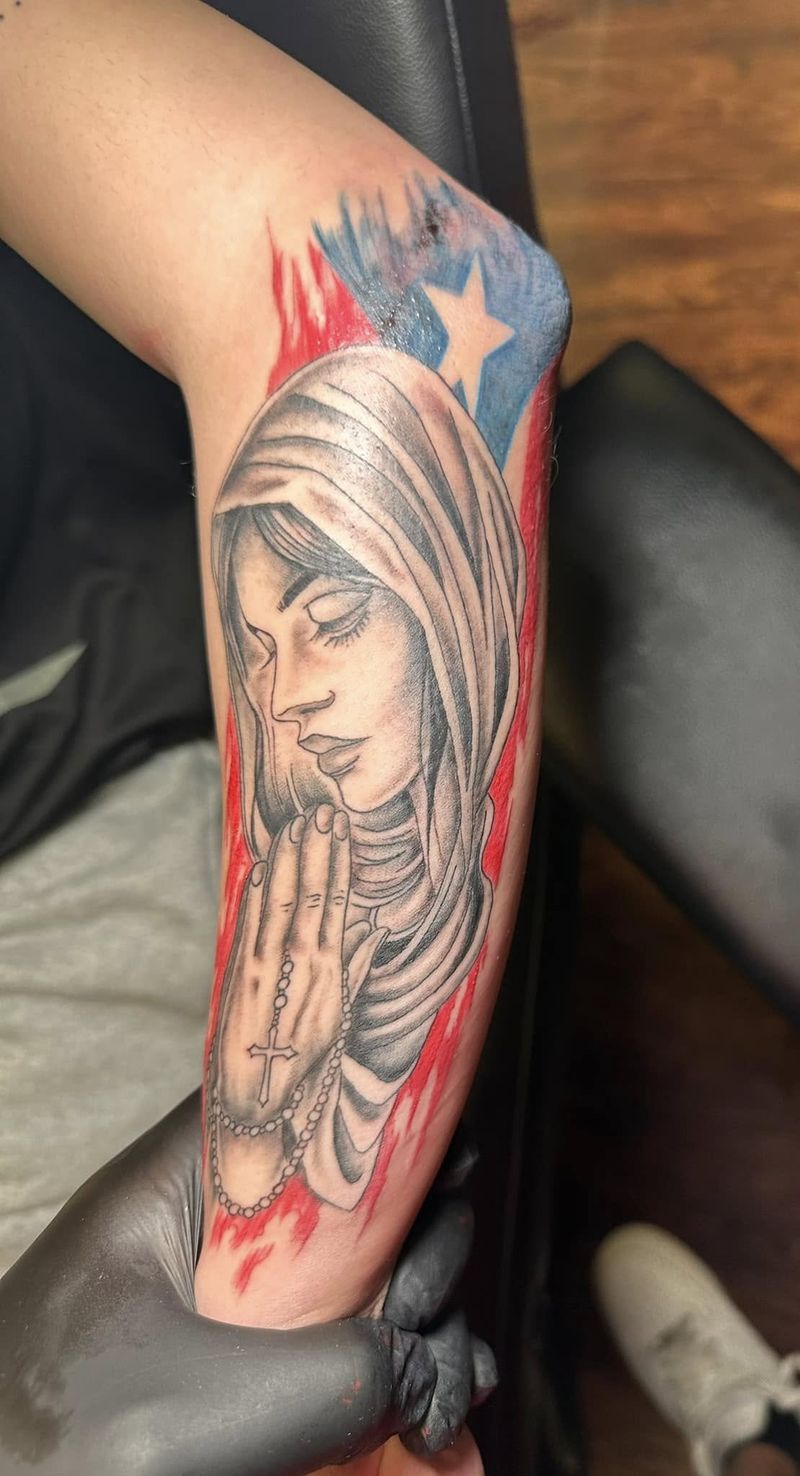 Tattoo of a veiled Madonna with praying hands holding a rosary, framed by red and blue flag details, on a forearm.