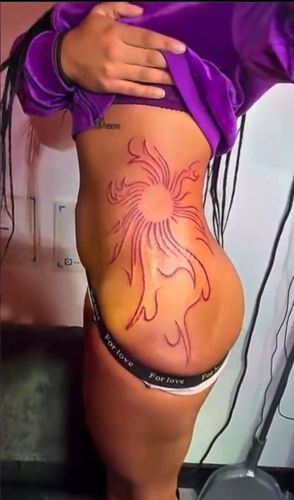 Red linework tattoo of a stylized sun with flowing rays on the side of the torso.