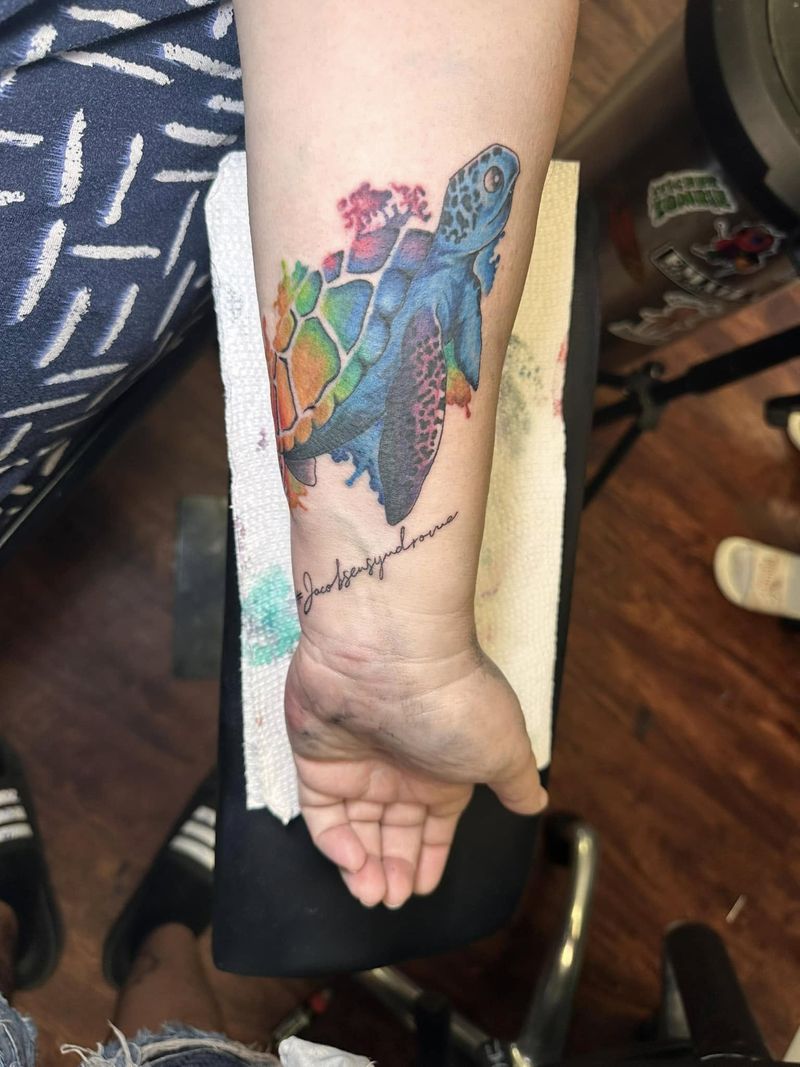 Color tattoo of a sea turtle in rainbow tones with a line of script beneath it, on the inner forearm near the wrist.