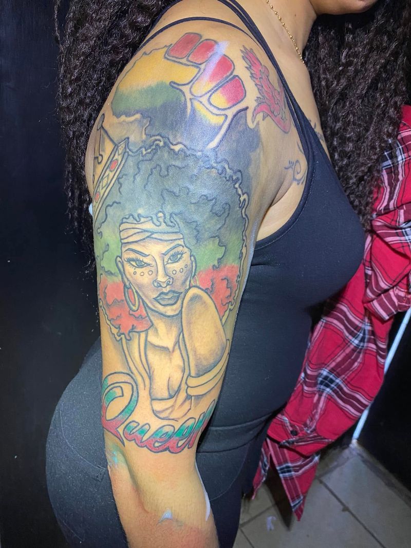Color tattoo of a woman with an afro beneath a raised fist on a red, yellow and green background, on an upper arm.